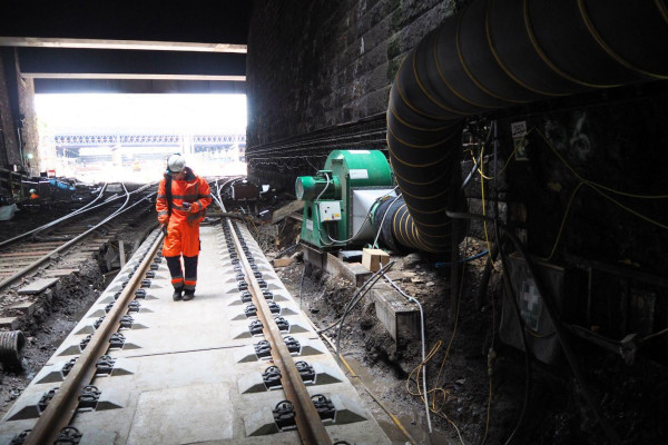 VENTEX Centrifugal Fan 800S to the right with a worker in orange hi-vis overalls walking on a railway track to the left. The tunnel opening is visible in the background_